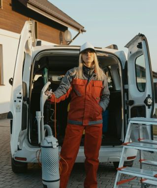 A cleaner in uniform stands beside a van with cleaning tools outside a modern house at sunset.
