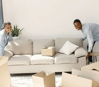 A cheerful couple moving furniture in a well-lit modern living room with cardboard boxes.